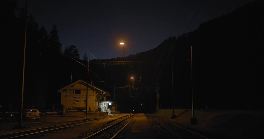 Train station emits light onto tracks at night with conifer forest in background under clear sky