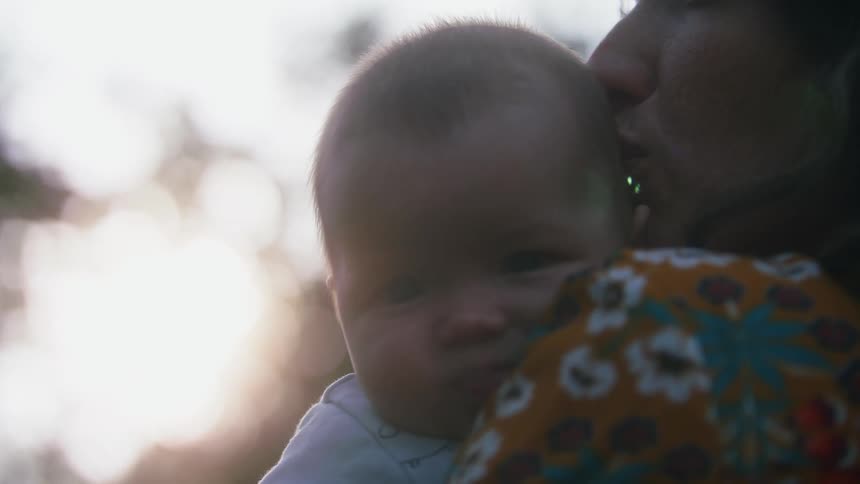 Mother with orange shirt with flowers holds baby boy looking over moms shoulder in the forest