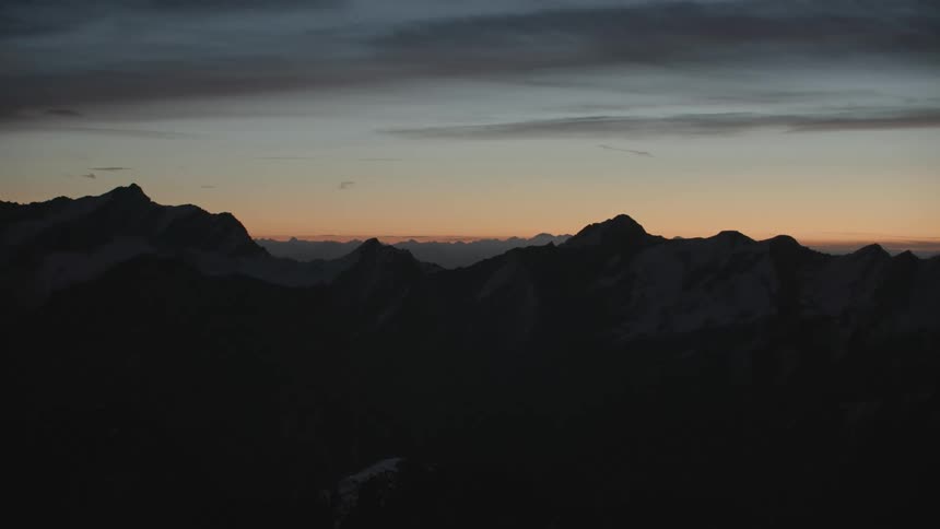 Dark shrouds mountain slopes and valley as snow covers mountain tops under red tinted sky