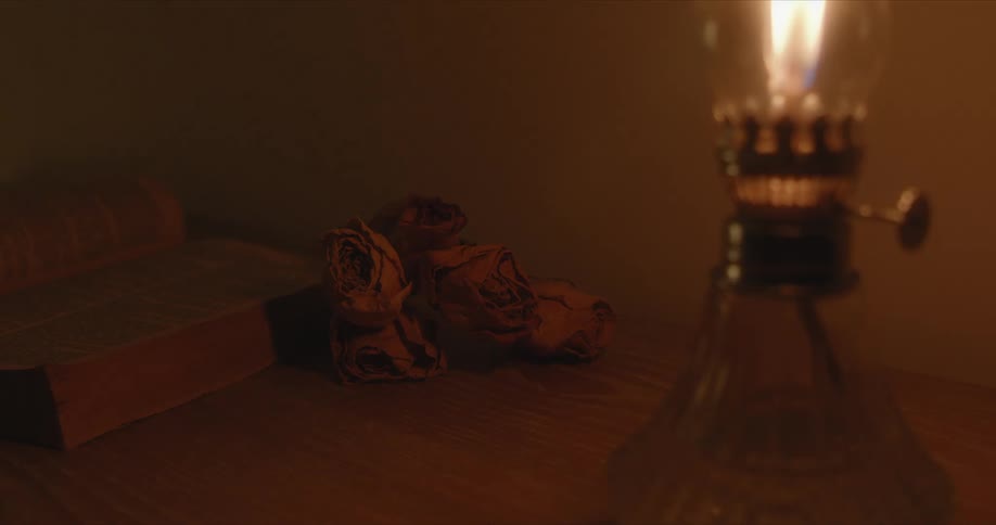 An oil lantern burns on a desk near dried flowers and an open book
