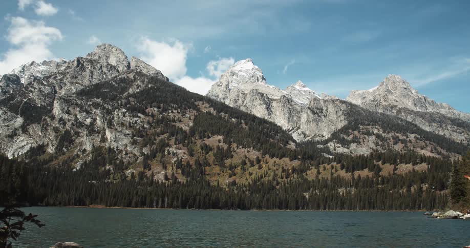 Clouds flow across sky casting shadows onto snowy mountain and nearby forest as people move along shore