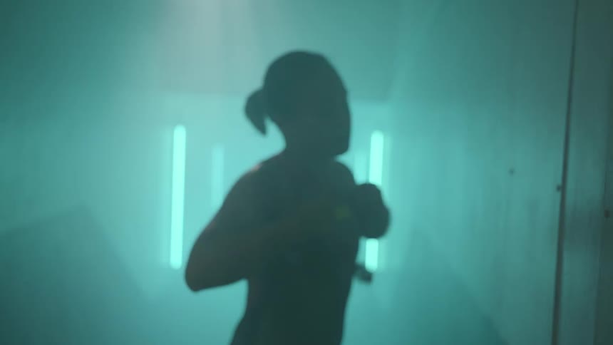 A teenage girl holding a swim cap and goggles adjusts her bathing suit as she paces in front of bright lights