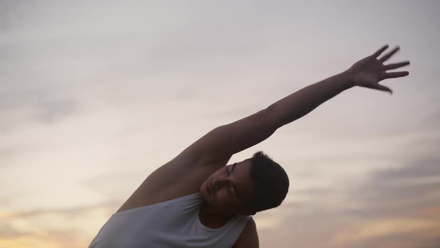 Man with white t shirt and black traning pants stretches his body and exercises in the field at dusk