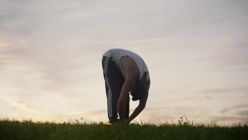 Young man wearing sports clothes stretches over the grass at evening