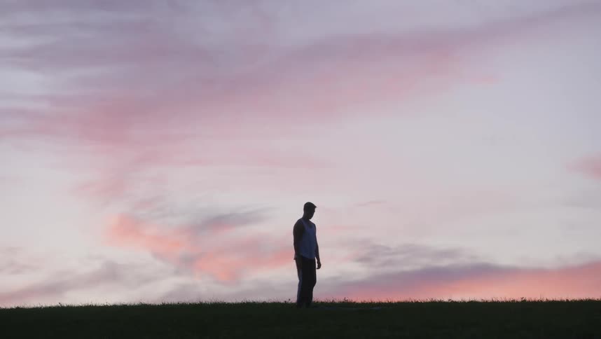 Man spread mat on grass and moves into yoga pose partly silhouetted against red tinted sky