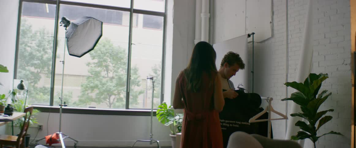 A man and a woman in a room with the woman handing the man a garment bag with a wall and window in the background