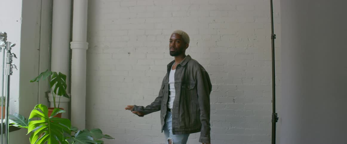 A man adjusts the collar of his jacket as he stands posing with a white wall backdrop