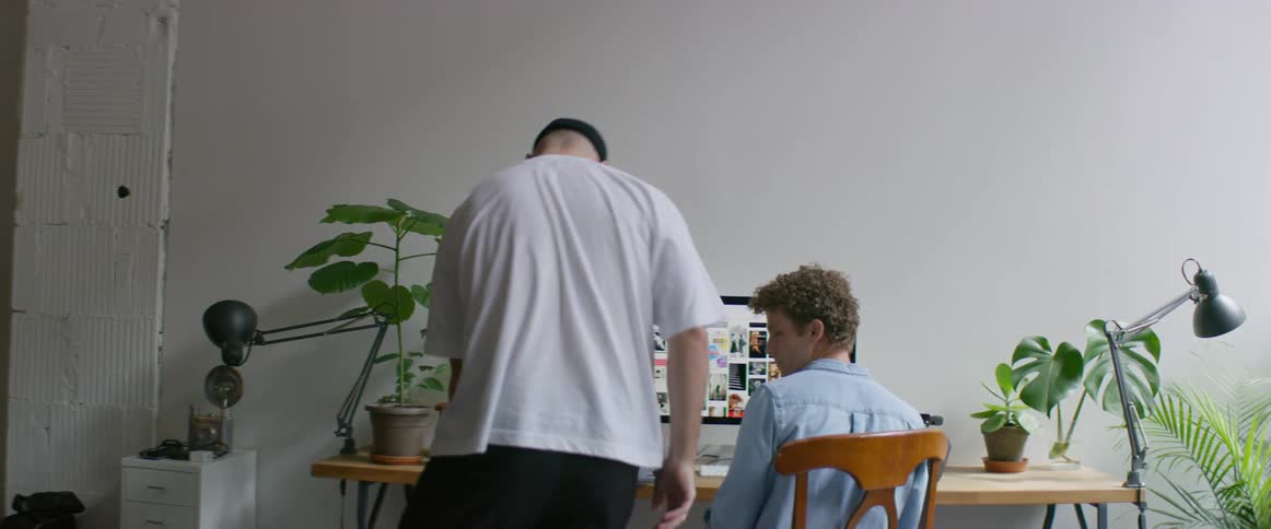 A man sits at his desk and looks at pictures on his laptop when another man comes in and they shake hands and he grabs a box and sits on it and looks at the computer also