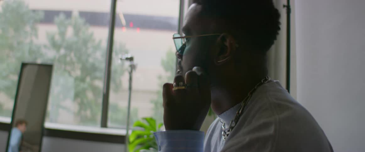 A man with sunglasses sitting in a room with a wall a mirror and a window in the background