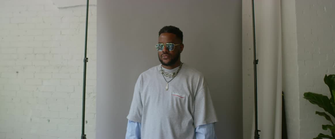 Young male model wearing sunglasses stands in front of a backing inside a room