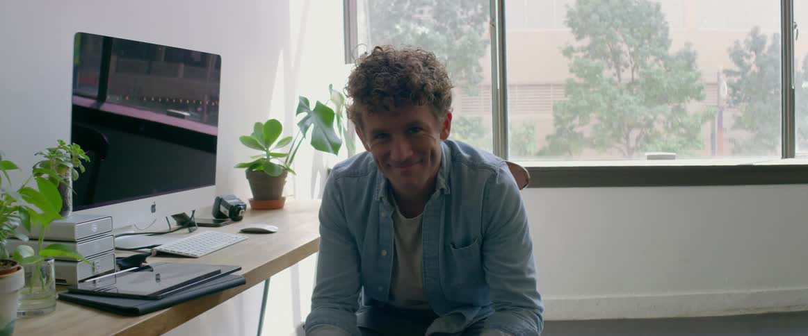 A man in a room sitting on a chair next to a desk and computer with a wall and window in the background