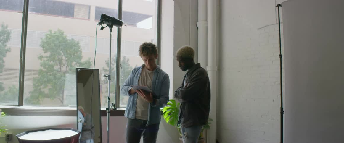 Photographer shares tablet screen with another man as he talks while they stand in photography studio