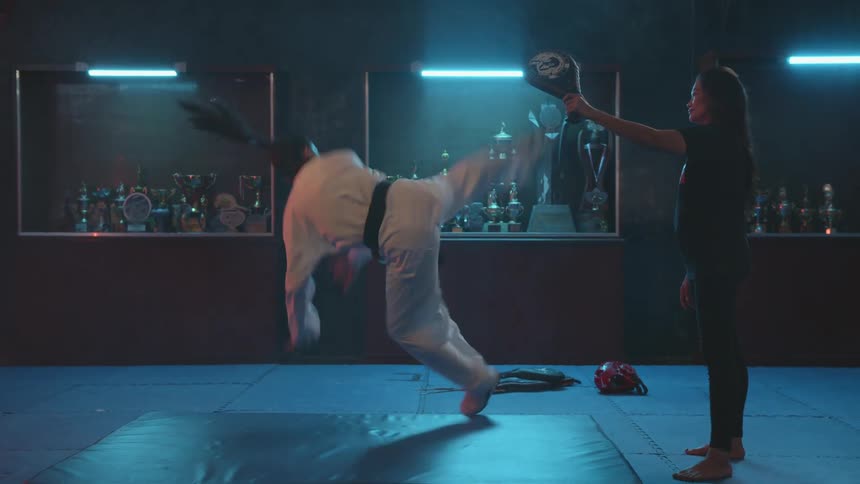 A woman in a room practicing martial arts with another woman with trophies shelves and a wall in the background