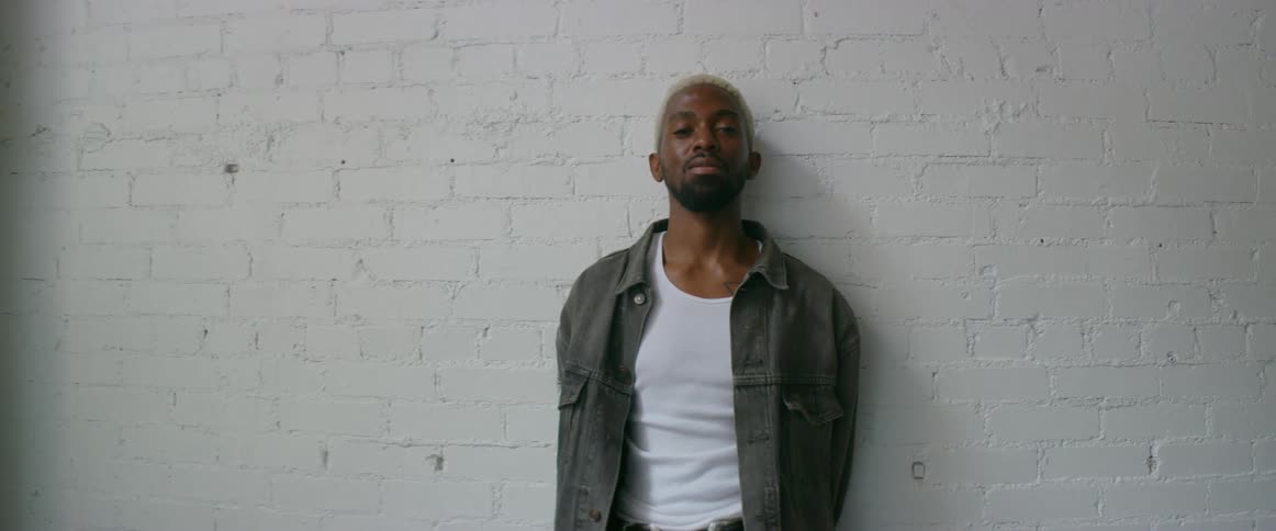 A black male model with bleached hair and facial hair poses in front of a white brick wall.