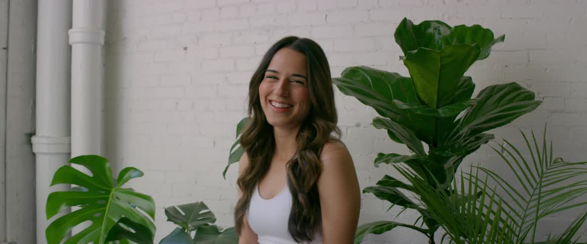 Woman sits near houseplants and white painted brick wall smiling and laughing
