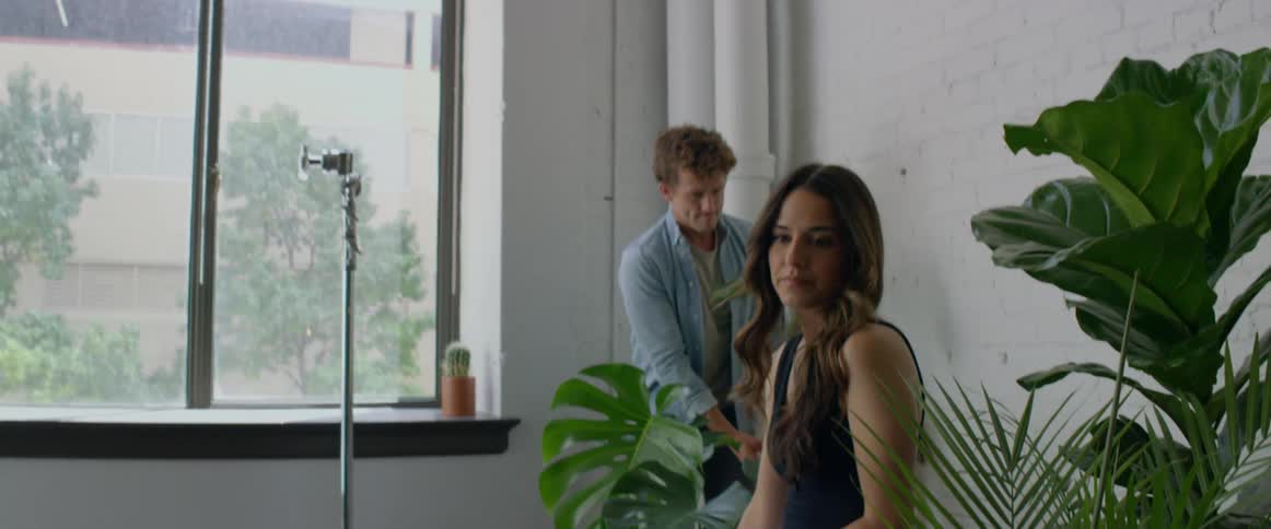 A man places plants around a woman posing on a chair