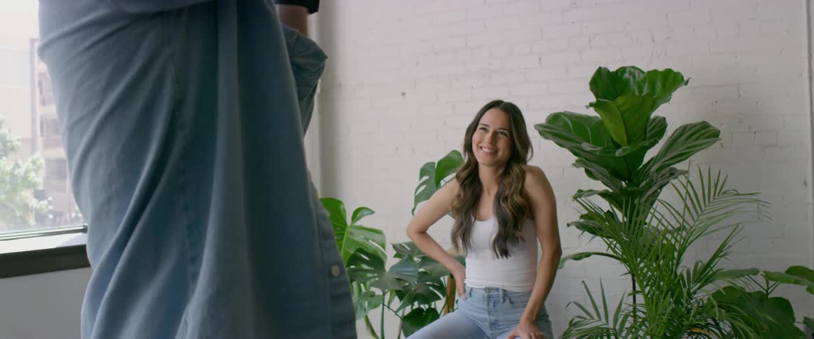 Male photographer with light blue shirt takes photographs of female model with brown wavy hair white top and jeans sitting in front of white wall between plants in the studio