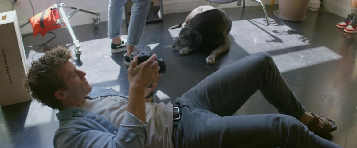 Photographer lies on back taking picture of model in photography studio as dog sleeps in shadow near sunlight coming from window