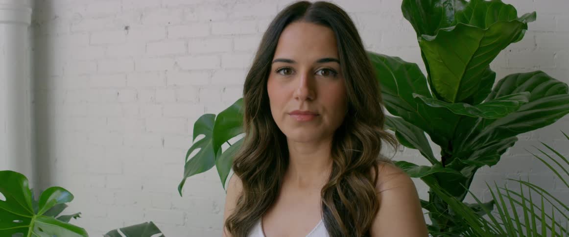 Woman poses against green houseplants and white painted brick wall
