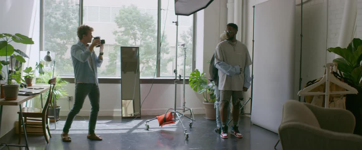 A man with a camera in a room taking pictures of two people with a wall and window in the background
