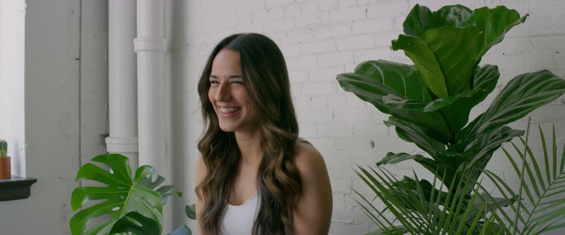 Woman with long wavy brown hair and white top laughs and smiles between green plants in front of a white wall