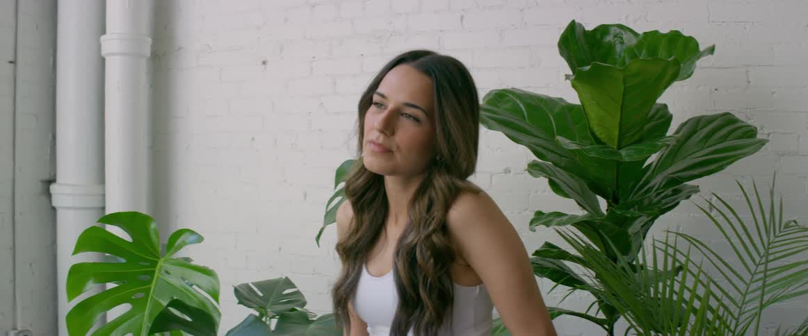 A model poses before a large house plant while being photographed by a photographer indoors during the day