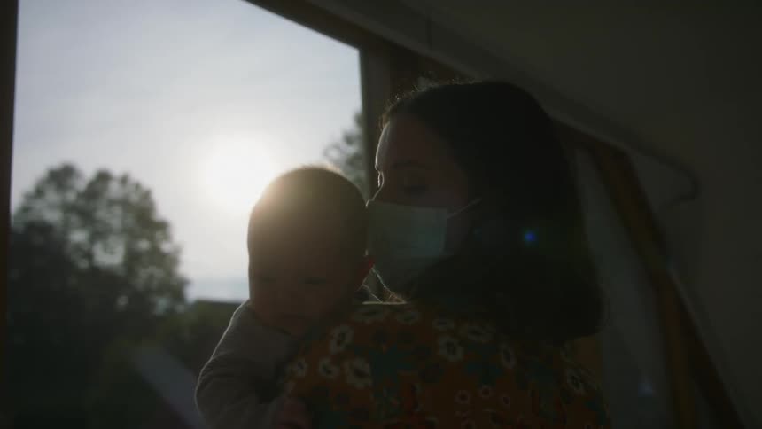 Mother wearing face mask hold baby while standing near window