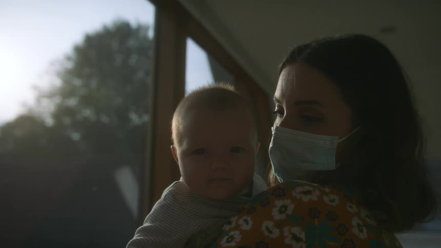 Woman wearing face mask holds baby while standing in home near window