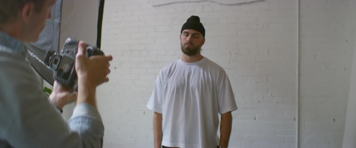 Man wearing white shirt and black beanie modeling for the camera in front of a white brick wall
