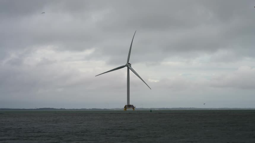 A large windmill located in a large body of water is spinning on an overcast and dreary day