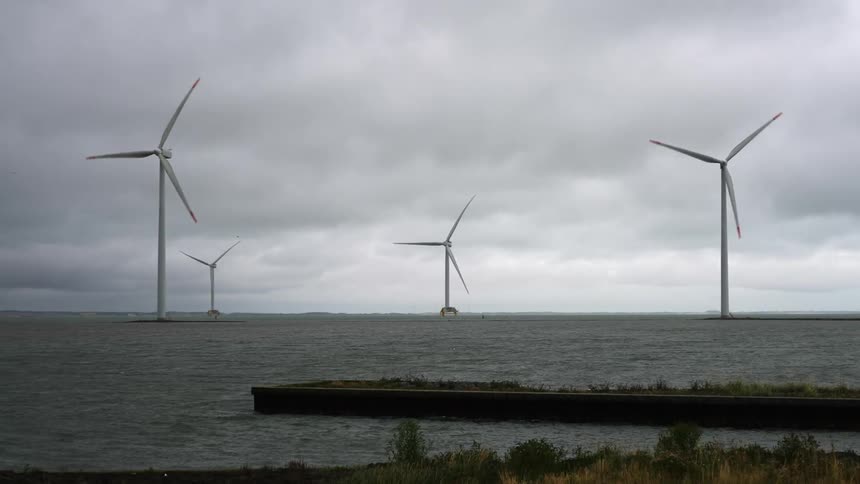 Wind turbines rotate on poles in rippling water under overcast sky as plants wave in wind on shore