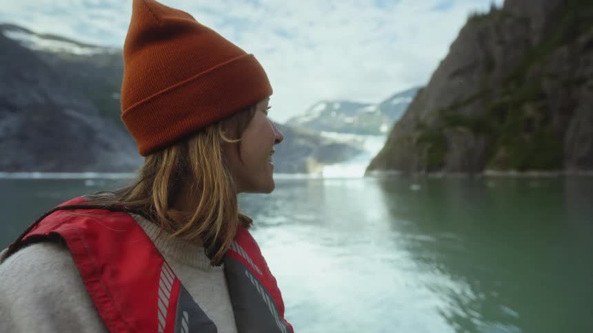 Woman wearing knit hat looks across reflective water gesturing to cliff and glacier