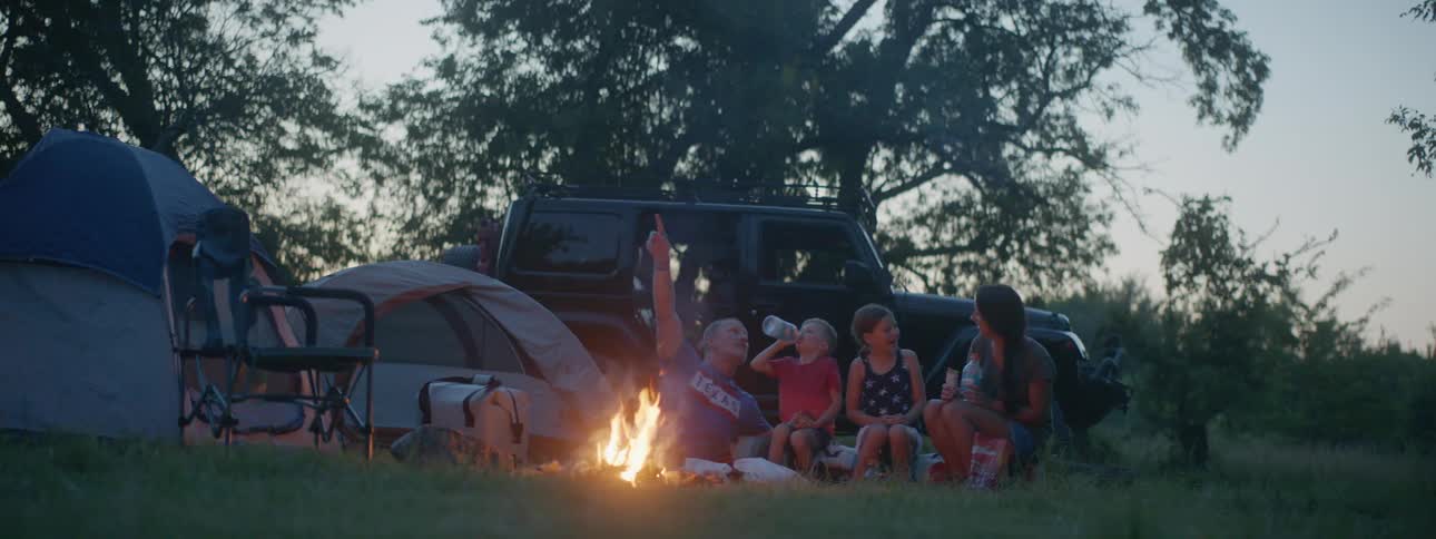 A man a woman a boy and a girl in front of a campfire next to tents with a vehicle trees the sky and clouds in the background