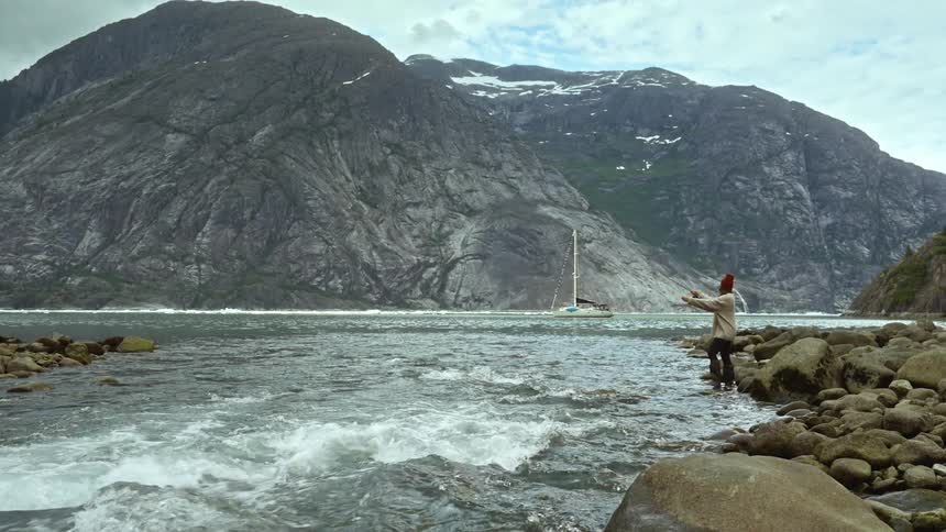 Woman with red beanie brown coat and black pants fly fishes into river next to high mountains