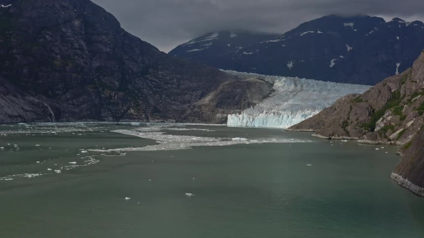 Sea ice floats on water near vegetation dotted mountains and glacier under overcast sky