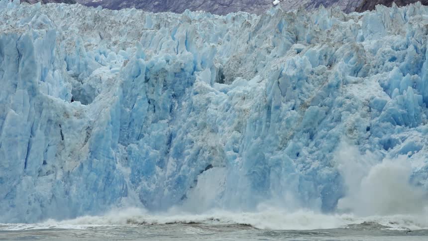 Birds fly past glacier as waves crash against shore creating spray and foam