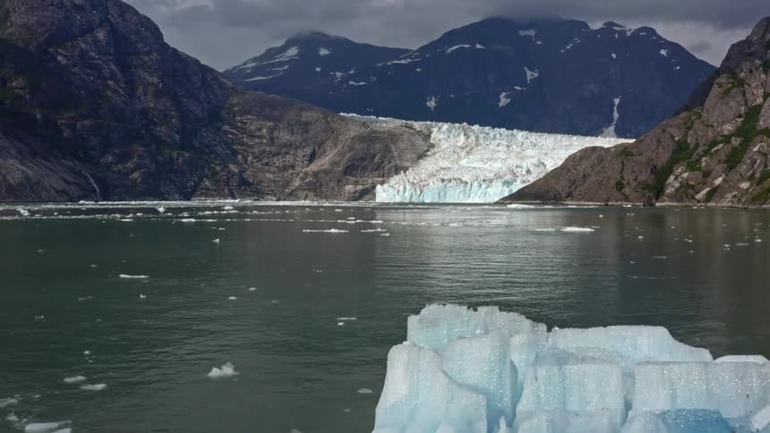 Ice floats across water from glacier sitting between mountains under overcast sky