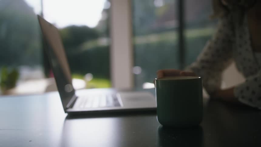 Young woman sitting at a desk at home puts down a hot beverage to work on a laptop