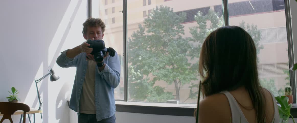 A man in a room with a camera taking a picture of a woman with a wall in the background