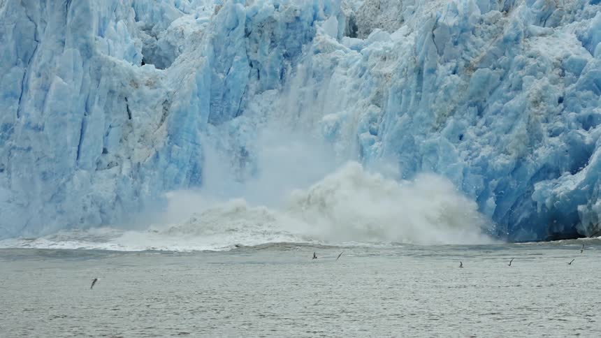 Birds are flying over water as snow and ice from an iceberg come crashing down