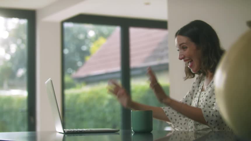 Woman sitting in the kitchen at home talking on a video call while drinking a hot beverage