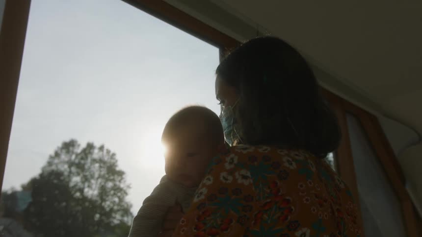 Mother with dark wavy hair orange blouse with flowers holds baby and the baby looks over mothers shoulder in front of windows