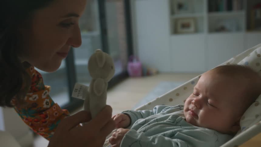 A woman in a room kissing a baby and holding a toy with a wall in the background