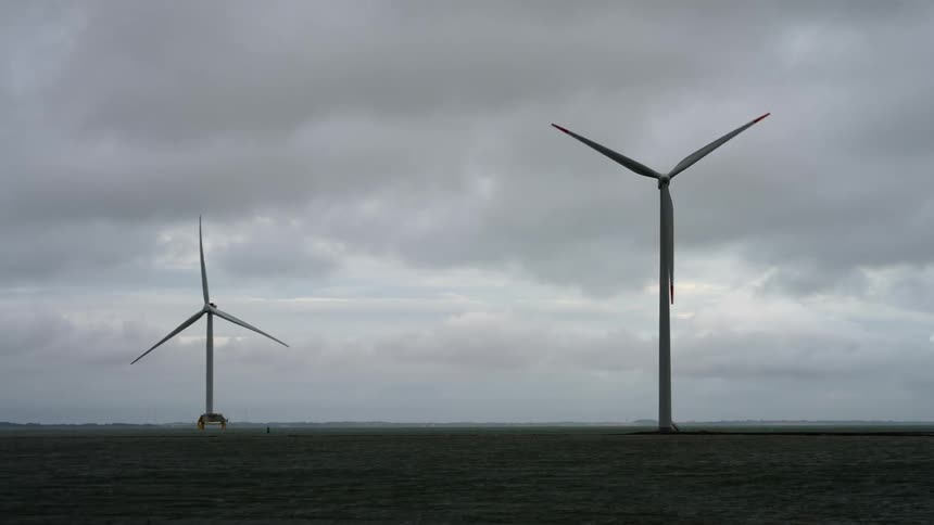 On a gray and cloudy day large windmills are spinning on a flat landscape