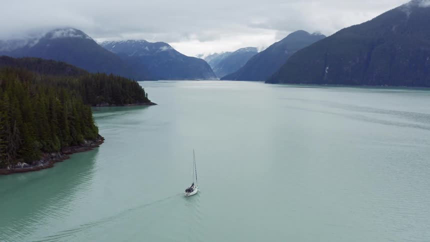 Sailboat with the sails moves across water passing conifer covered forest creating wake with mountains in background