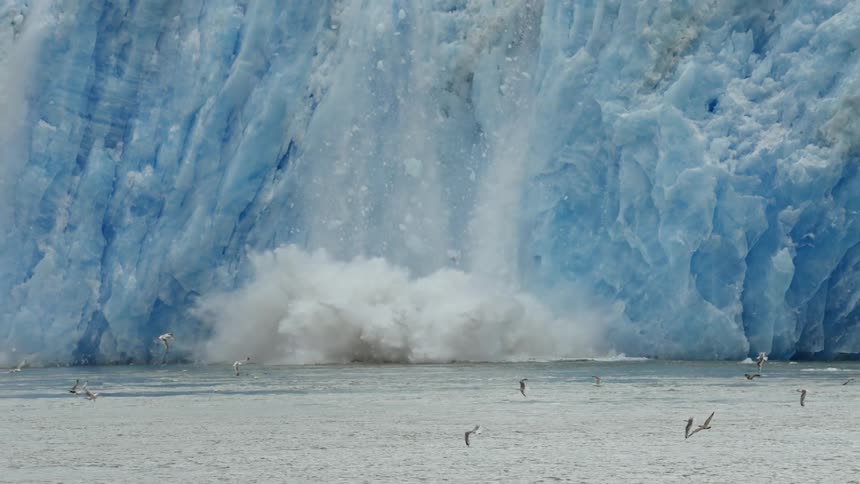 Birds are flying over water as snow and ice from an iceberg come crashing down