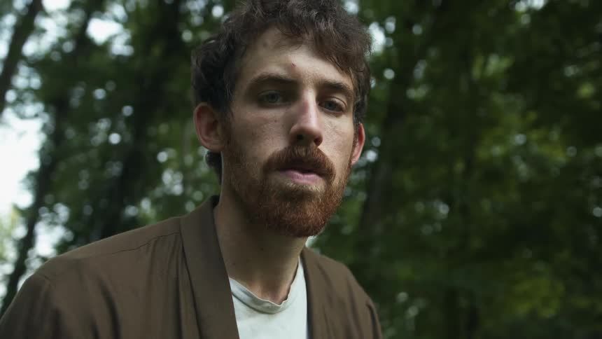 Man with red beard white and brown shirt stares down at camera in the forest