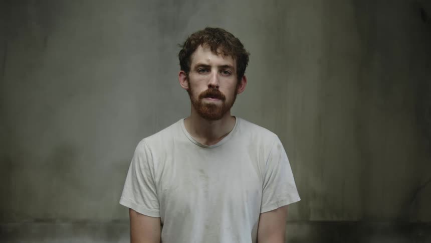 Red haired man wearing t shirt with beard and mustache stands staring as shadows and light flow across him and wall