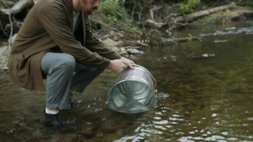 Man with red beard white and brown shirt blue pants and boots takes bucket of water from stream in the forest