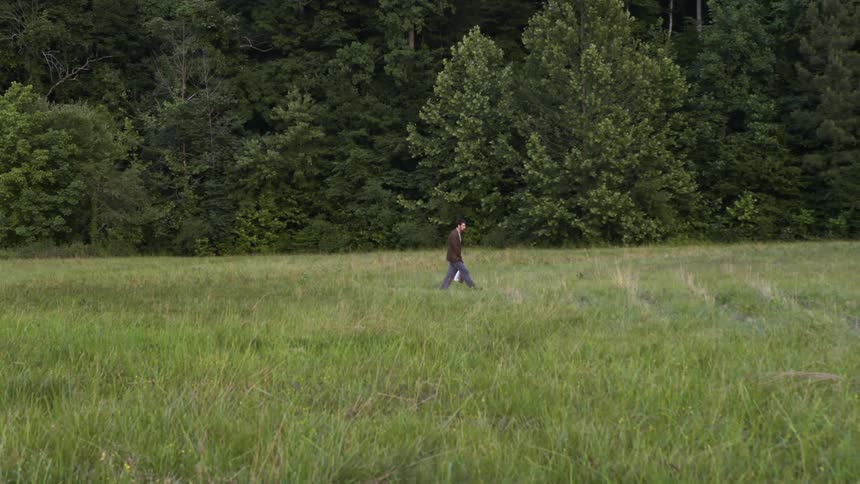 Man walks across grassy field paralleling woods while carrying bag